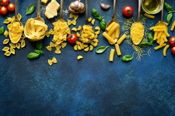 Assortment of pasta on a spoons with ingredients for making : tomato, parmesan chheese, garlic, basil and olive oil. Top view with copy space.