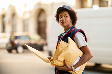 Black delivery woman with packages looking away on city street.