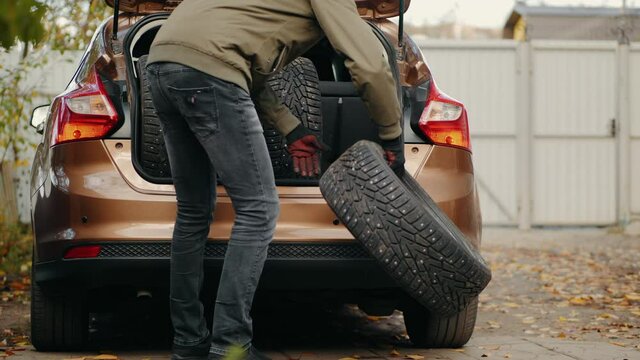 Rear view: A man unloads studded winter tires from a car trunk. Motorist changes car tires from summer to winter