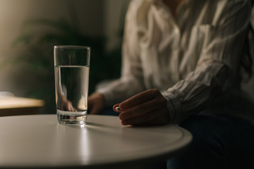 Sick woman holding a pill in her hand, close-up. There is a glass of water on the table in a dark room. The girl plans to take the pill.