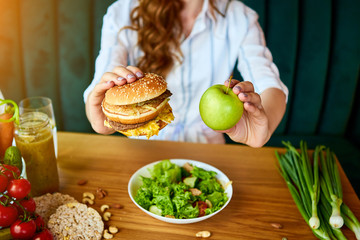 Beautiful young woman decides eating hamburger or apple in kitchen. Cheap junk food vs healthy diet