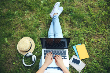 Young girl using laptop computer with headphones, smartphone and books on the grass in park