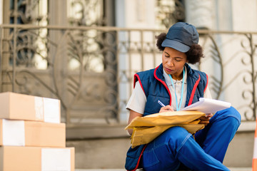 Black female courier taking notes going through delivery list. - obrazy, fototapety, plakaty