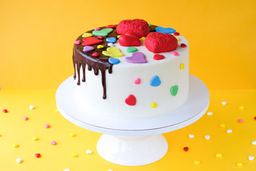 White cake decorated with colorful hearts, confectionery sprinkles and drenched in chocolate on a yellow background.