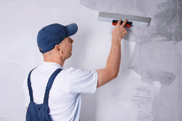 Worker in blue overalls and cap plastering a wall with finishing putty using a putty knife. Repair work and construction concept. Close-up