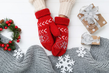 Hands in knitted mittens with grey scarf, snowflakes and gift boxes on white wooden table