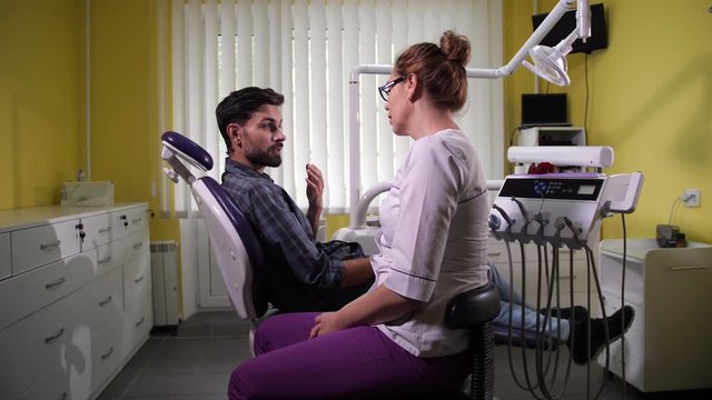 Female stomatologist talking with male client sitting in chair during consultation in dental office. Patient telling woman dentist about his teeth problems before primary examination