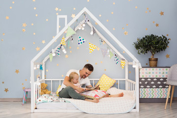 Adorable little girl and her young dad are reading a book while sitting in decorated house bed at...