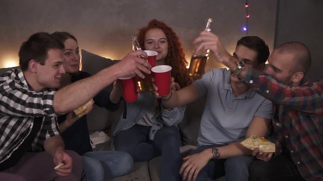 Portrait of diverse group of friends have delivered pizza celebrating - happily drinking beer, cheers. Having fun party in cozy room decorated with garland lights in a company of close friends