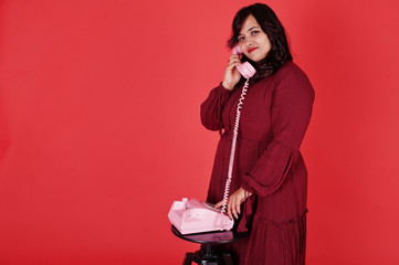 Attractive south asian woman in deep red gown dress posed at studio on pink background and use old vintage telephone.