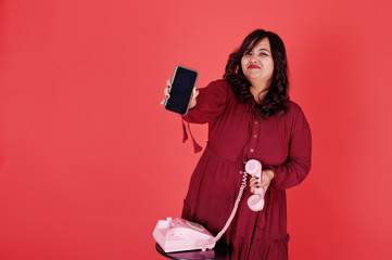 Attractive south asian woman in deep red gown dress posed at studio on pink background and use old vintage telephone with modern mobile phone.