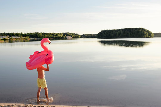 Young man with flamingo pool float walking at lakeshore