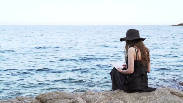 Caucasian girl in black hat and black dress sitting by the ocean and reading book. Summer chill at ocean seashore. Education and leisure by the sea shore.  Blue water and sunset light, fancy dress.