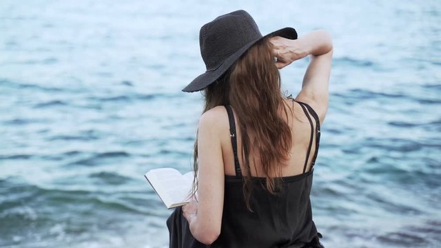 Caucasian girl in black hat and black dress sitting by the ocean and reading book. Summer chill at ocean seashore. Education and leisure by the sea shore.  Blue water and sunset light, fancy dress.