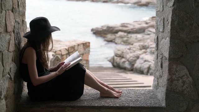A girl in a black hat and black silk dress sits in the niche of a large window in a fishing hut on the seashore and reads a book. Ocean view at a background. Studying outdoors, reading a novel.
