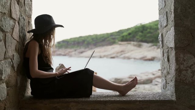 A girl in a black hat and black silk dress sits in the niche of a large window in a fishing hut on the seashore and working on laptop. Ocean view at a background. Studying outdoors, texting. Freelance