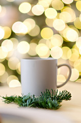 New year greeting card. On the wooden table is a gray cup of hot chocolate, decorated with a Christmas tree branch. Blurred bright yellow lights bokeh background. Festive drinks. Copy space. Texture