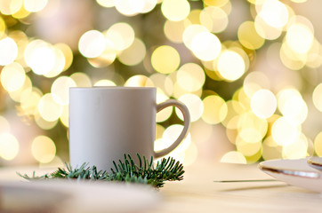 New year greeting card. On the wooden table is a gray cup of hot chocolate, decorated with a Christmas tree branch. Blurred bright yellow lights bokeh background. Festive drinks. Copy space. Texture