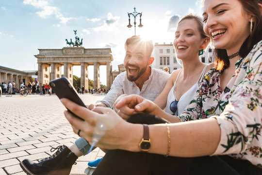 Three laughing friends sitting near Brandenburger Tor looking at smartphone, Berlin, Germany