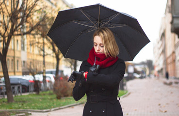 A woman looks at her wrist, smart watch, awaits a meeting. Girl on a cold day, dressed in seasonal clothes with an umbrella in her hands.