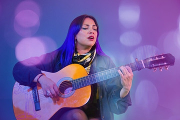 Obraz na płótnie Canvas Latin woman playing an acoustic guitar on purple background with drained blue and light sparkles