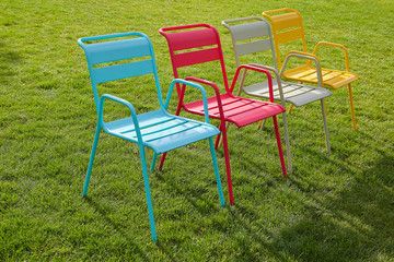 Four multi-colored chairs stand on a green lawn on a summer evening. Modern metal chairs are...