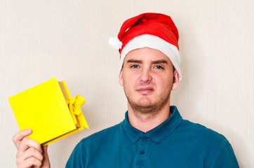 upset, displeased handsome guy in santa hat with disgust holds in his hand a box with gift. young...