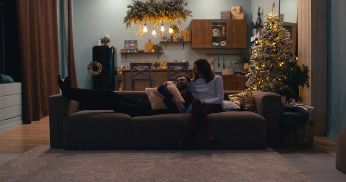Caucasian couple sitting together on a sofa and watching TV at home during Christmas holidays, decorated apartment interior. 4K UHD RAW graded footage