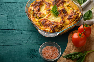 Ready Italian lasagna, on a green wooden background with basil, tomatoes. Culinary background, Italian recipe book, delicious food. With space for design, top view.