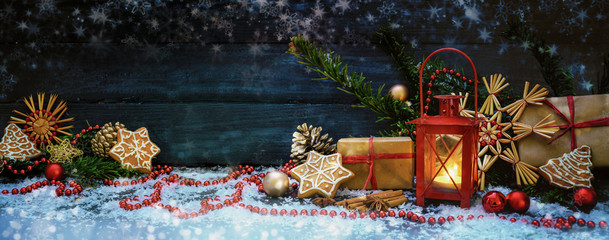 Wide Christmas background wit red candle light lantern, gifts, baubles and decoration, stars and snowflakes against a dark blue wooden background, panoramic format with copy space