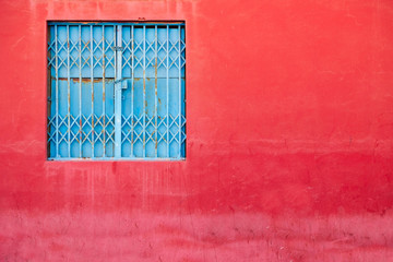 Square blue closed and barred window in vibrant red grungy wall.