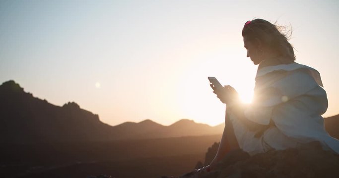 Young dreaming woman using smartphone on mountain top at sunset in wonderful nature. Spectacular refreshing scenery of volcanic mountains in Teide, Tenerife.
