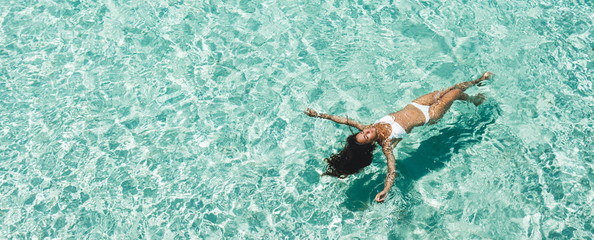 Woman in white bikini lying on transparent turquoise water surface on beach. Travel and vacations concept. Tropical background with empty space - obrazy, fototapety, plakaty