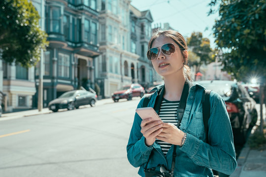 vintage style portrait of young girl backpacker sightseeing tour alone in modern urban. beautiful lady tourist in sunglasses holding cellphone walking in San Francisco painted ladies road on sunshine