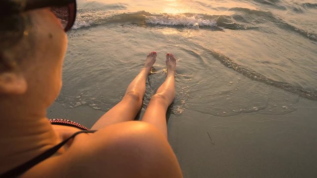 Young woman legs relaxing on a beach among sea waves in a sunset