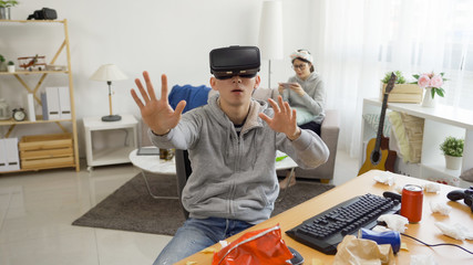 Young asian student boy wearing virtual reality glasses at home on summer break. asian male people having fun with new technology vr headset goggles. girlfriend in back relax on couch using cellphone