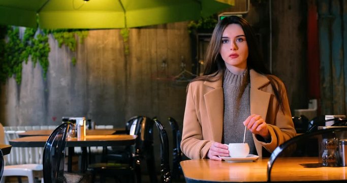 a young woman sits in a cafe and drinks a hot drink from a white cup. she's wearing an autumn coat and a warm sweater. the concept of a romantic evening and the relations. 4k proress 50p
