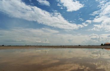 Obraz premium View of salf field under bright blue sky, white fluffy clouds casting reflections over surface of sea water. Salt farming in Thailand.