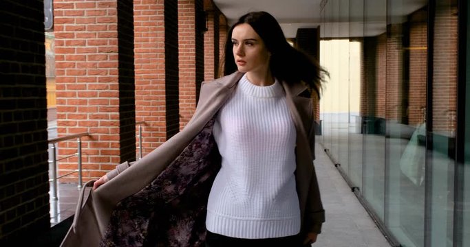 a girl in an autumn coat walks next to an office building with brick walls and a storefront. the girl waves her coat unbuttoned and sometimes there is sunlight