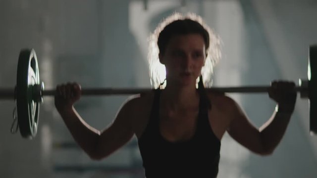 A beautiful sportswoman in black sportswear does a squat with a barbell load on her back in a dark hall. The concept of strength and motivation