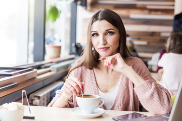 Attractive young woman sits at a table in a cafe with a cup of coffee and mixes it with a spoon