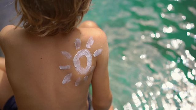 Slowmotion shot of a little boy with a sun drawn by a sunscreen cream on his shoulder splash water in a swimming pool. Sun protection concept