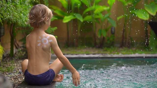 Slowmotion shot of a little boy with a sun drawn by a sunscreen cream on his shoulder splash water in a swimming pool. Sun protection concept