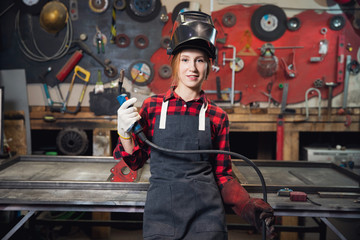 Young woman welder smiles and holds protective mask in hand on background industrial workplace or...