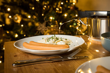 Wiener Würstchen und Kartoffelsalat als typisches Weihnachtsessen zu Heiligabend in Deutschland am Weihnachtsbaum mit Weihnachtsdekoration und Lichterketten