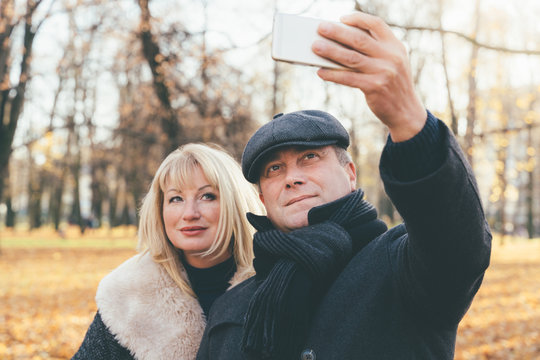 Happy blonde mature woman and beautiful middle-aged brunette take selfie on mobile phone. Loving couple of 45-50 years old walks in autumn park in warm clothes, in coat and enjoys life