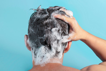 Male hand washes his head with shampoo and foam on a blue background, back view