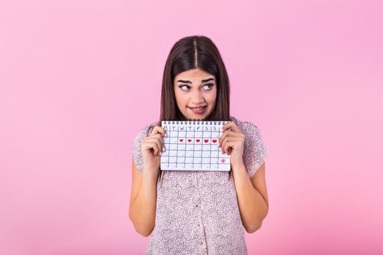 Portrait of embarrassed cute girl holding menstrual calendar with drawn hearts for period and looking at camera isolated over pink background