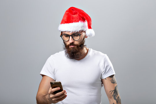 Christmas, holidays, technology and people concept - handsome bearded man in santa hat taking selfie picture with smartphone on grey background