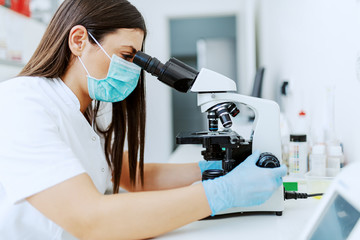 Side view of female lab assistant with mask and rubber gloves using microscope for examining sample.